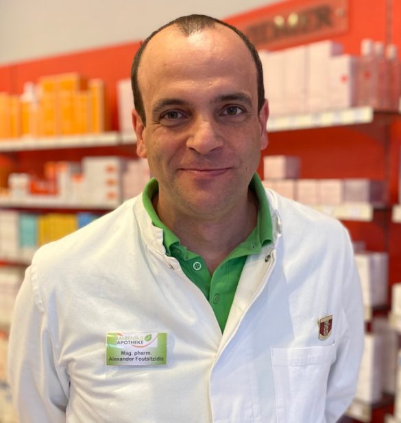 Ein Mann in einem weißen Laborkittel steht lächelnd in der Laurentius Apotheke Imst / Pitztal Apotheke. Im Hintergrund sind Regale mit Medikamentenschachteln zu sehen.