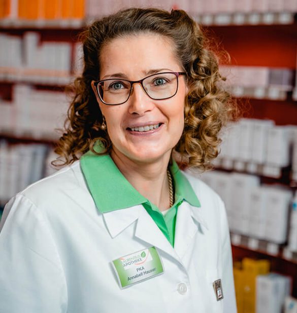 Eine Frau mit lockigem Haar und Brille, die einen weißen Laborkittel und ein Namensschild trägt, steht vor Regalen, die mit Apothekenartikeln gefüllt sind, in der Laurentius Apotheke Imst / Pitztal Apotheke.
