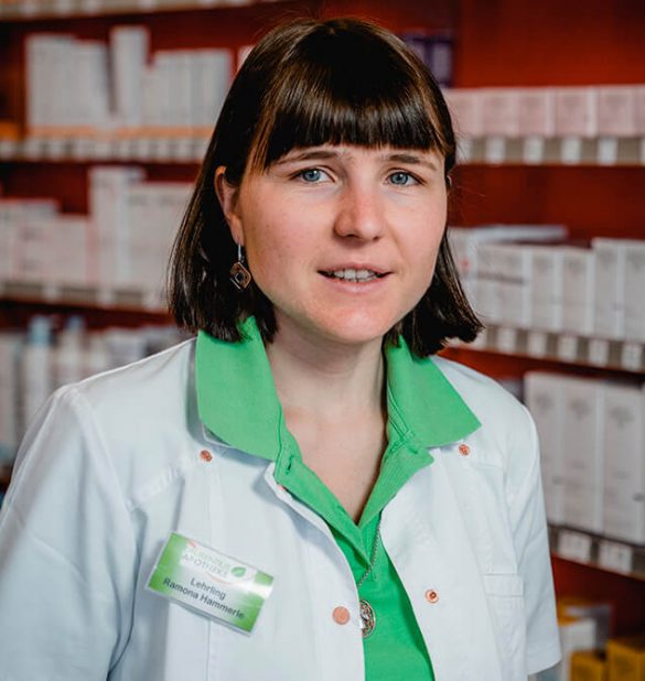 Eine Frau mit glattem braunem Haar, die einen weißen Laborkittel der Laurentius Apotheke Imst / Pitztal Apotheke und ein grünes Hemd trägt, steht vor Regalen, die mit verpackten Produkten gefüllt sind.